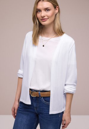 Jonge vrouw met blond haar, gekleed in een wit vest, wit overhemd, blauwe spijkerbroek, een beige riem en gelaagde gouden kettingen tegen een beige achtergrond.