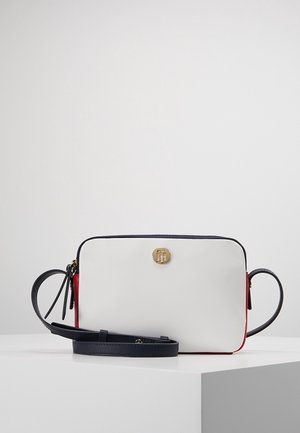 Sac bandoulière rectangulaire blanc avec bords noirs, panneaux latéraux rouges, un logo circulaire doré et une sangle noire réglable posé sur une surface blanche.