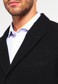 Man met een stoppelbaard in een zwart colbert over een wit overhemd met een lichtblauw ruitpatroon, met een open kraag en knoopjes aan de bovenkant.