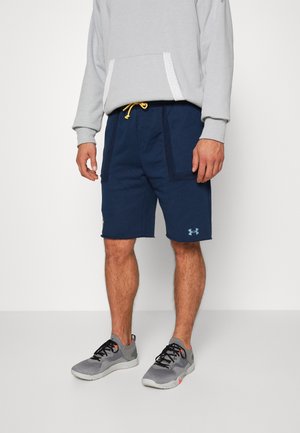 Man wearing navy blue sports shorts with yellow drawstring, grey hoodie, and grey athletic shoes with black laces on plain white background.