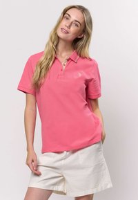 Ružové tričko polo s krátkymi rukávmi a kontrastným béžovým golierom, s malým vyšívaným logom na hrudi. Biele šortky s vreckami.