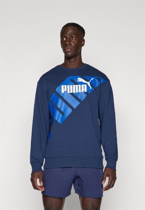 Granatowa bluza z miękkiego materiału z dużym białym logo "PUMA" oraz ukośnymi niebieskimi akcentami. Żebrowane mankiety i dół dopełniają projekt.