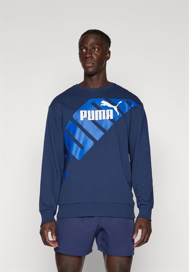 Felpa blu navy in morbido tessuto con un grande logo bianco "PUMA" e accenti diagonali blu. I polsini e l'orlo a coste completano il design.