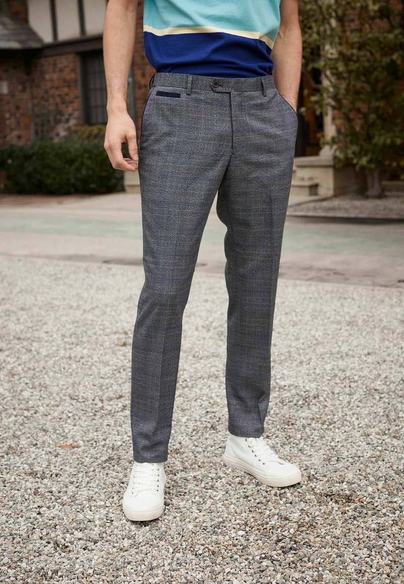 Pantalons à carreaux gris sur mesure avec un devant plat, des poches latérales et une coupe fuselée, associés à des baskets blanches sur une surface en gravier.