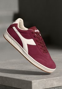 Sneakers din suede cărămizii cu accente albe, având un design cu șireturi, talpă din cauciuc și logo pe lateral. Materiale texturate și siluetă casual.