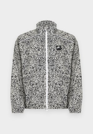 Veste légère Nike avec un motif noir et blanc tacheté, col montant, fermeture éclair complète à l'avant, et un petit écusson logo sur la poitrine.