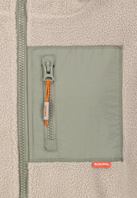 Fleece-Jacke mit einem beigen strukturierten Oberteil und einer blassen grünen Tasche mit einem orangefarbenen Reißverschluss, gebrandet mit "SUBLEVEL" auf einem Etikett.