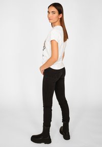 Kvinde med langt brunt hår iført en hvid grafisk T-shirt, sorte stramme jeans og sorte kraftige ankelsko, stående mod en ensfarvet baggrund.