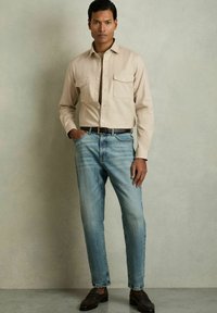Cămașă lungă de culoare bej deschis, cu două buzunare pe piept, asortată cu blugi drepți bleu decolorat și pantofi loafers maro, pe un fundal neutru.
