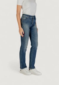 Mujer vistiendo unos pantalones vaqueros rectos de un azul desgastado, una camiseta blanca, deportivas blancas y con esmalte de uñas rojo, de pie sobre un fondo blanco.