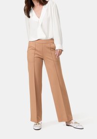 Blouse blanche à col en V avec manches longues, associée à un pantalon large marron clair; le mannequin porte des chaussures blanches à enfiler avec des accents dorés.