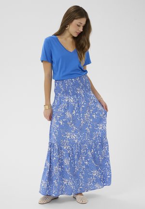 Frau trägt ein blaues V-Ausschnitt-T-Shirt, einen blau-weißen Blumen-Maxi-Rock, goldene Creolen und beige gewebte Slipper, posiert vor weißem Hintergrund.