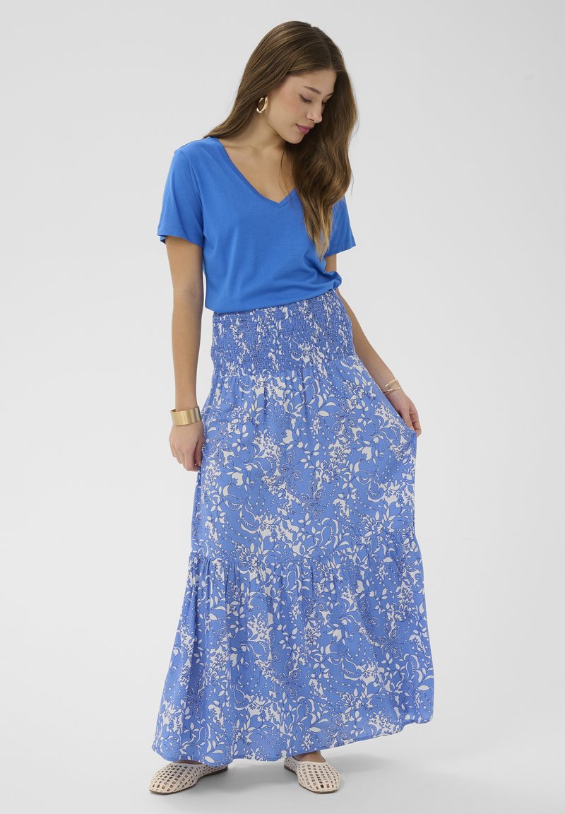 Femme portant un t-shirt bleu à col en V, une jupe maxi fleurie bleue et blanche, des boucles d'oreilles créoles en or et des chaussures beige tissées à enfiler, posant devant un fond blanc.