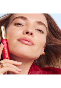 Czerwony serum do oczu NUXE Eye Lighter w eleganckiej tubce ze złotą nakrętką, trzymane przez modelkę o gładkiej skórze i złotych kolczykach w kształcie kół, na jasnoniebieskim tle.