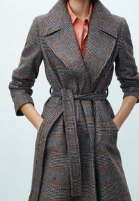 Grå ternet trenchcoat med bælte, der har en stor krave, flagrende ærmer og orange accenter i mønsteret. Stoffet ser tekstureret ud.