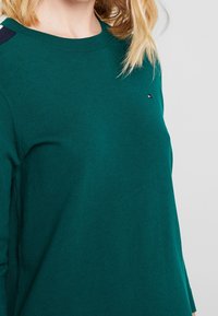 Grüner gestrickter Pullover mit rundem Halsausschnitt und strukturiertem Material. Er hat marinefarbene Akzente an den Schultern und ein kleines Logo auf der Brust.