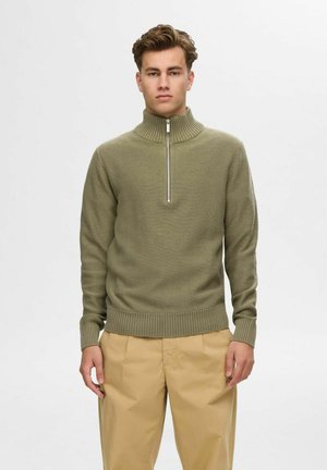 Pulóver de punto acanalado en verde oliva, con un alto cuello y media cremallera. Presenta mangas largas y cintura ajustada. Combinado con pantalones beige.