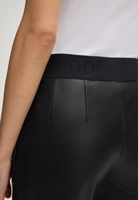 Close-up of a person wearing black faux leather pants with a wide black waistband featuring embossed "JOOP!" text, paired with a white top.