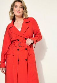 Rode trenchcoat met een nauwsluitend ontwerp, dubbele knopen, een ceintuur in de taille en twee zijzakken. Gemaakt van een gladde, lichte stof.