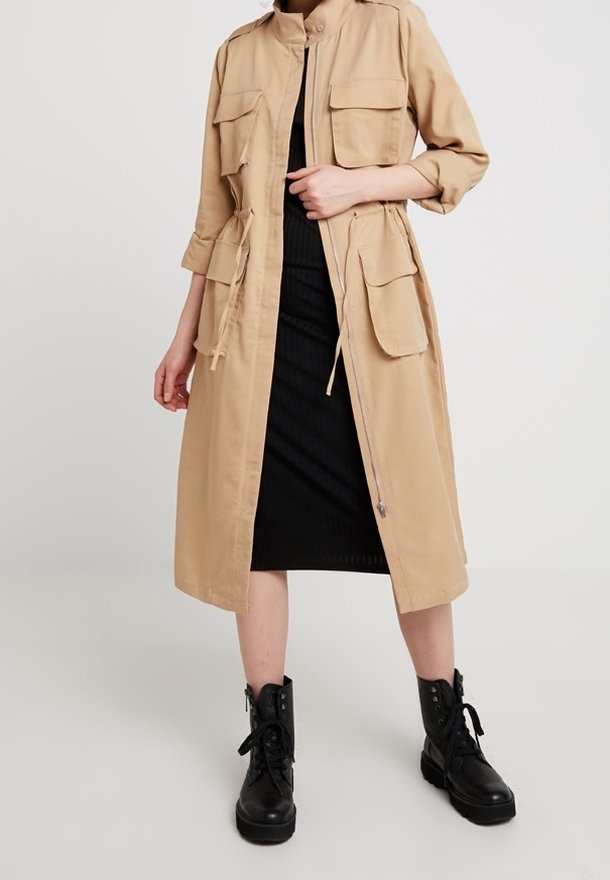 Veste utilitaire beige longueur genou avec quatre poches avant, un col montant et une fermeture éclair, portée sur une robe noire côtelée et des bottes noires.