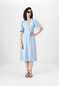 Rochie midi de culoare bleu deschis, confecționată dintr-un material moale, cu mâneci scurte tip puff, decolteu în V și închidere cu butoane în față. Sandale negre cu bretele.