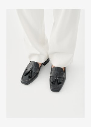 Mustad nahast mokassiinid tuttidega, ruudukujulise varbaosaga ja dekoratiivse õmblusega, kombineeritud valgete laiade pükstega.