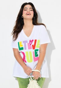 T-shirt blanc à col en v arborant un texte en blocs colorés avec les mots "LAISSEZ L'AMOUR RÉGNER" en rose, vert, jaune et violet. Manches courtes, tissu doux.