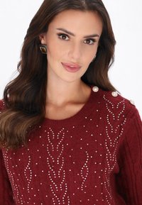 Czerwony sweter z dzianiny o fakturowanym wzorze, ozdobiony cyrkoniami i perłowymi detalami na ramionach. Posiada okrągły dekolt.