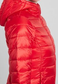 Czerwona kurtka puchowa o błyszczącym wykończeniu, z kapturem, pikowanym wzorem i bocznymi kieszeniami. Materiał wydaje się lekki i ocieplający.