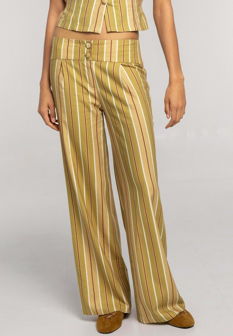 Mujer con pantalones de pierna ancha y talle alto en color mostaza con rayas verticales blancas y rojas, combinados con un top corto a juego y zapatos marrones.