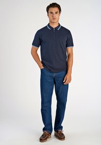 Marineblå polo med dragkedja och kontrasterande blå detaljer, i kombination med sten tvättade blå jeans och bruna båt skor. Enhetlig bakgrund.