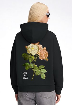 Eine Person mit Sonnenbrille trägt einen schwarzen Hoodie mit großem Rosenmotiv und den Worten "LOVE & ROSES" auf dem Rücken, kombiniert mit grauen Jeans.
