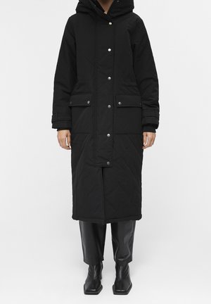 Personne portant un long manteau d'hiver noir à capuche avec des boutons-pression, des poches à rabat, ainsi qu'un pantalon en cuir noir et des bottes visibles sous le manteau.