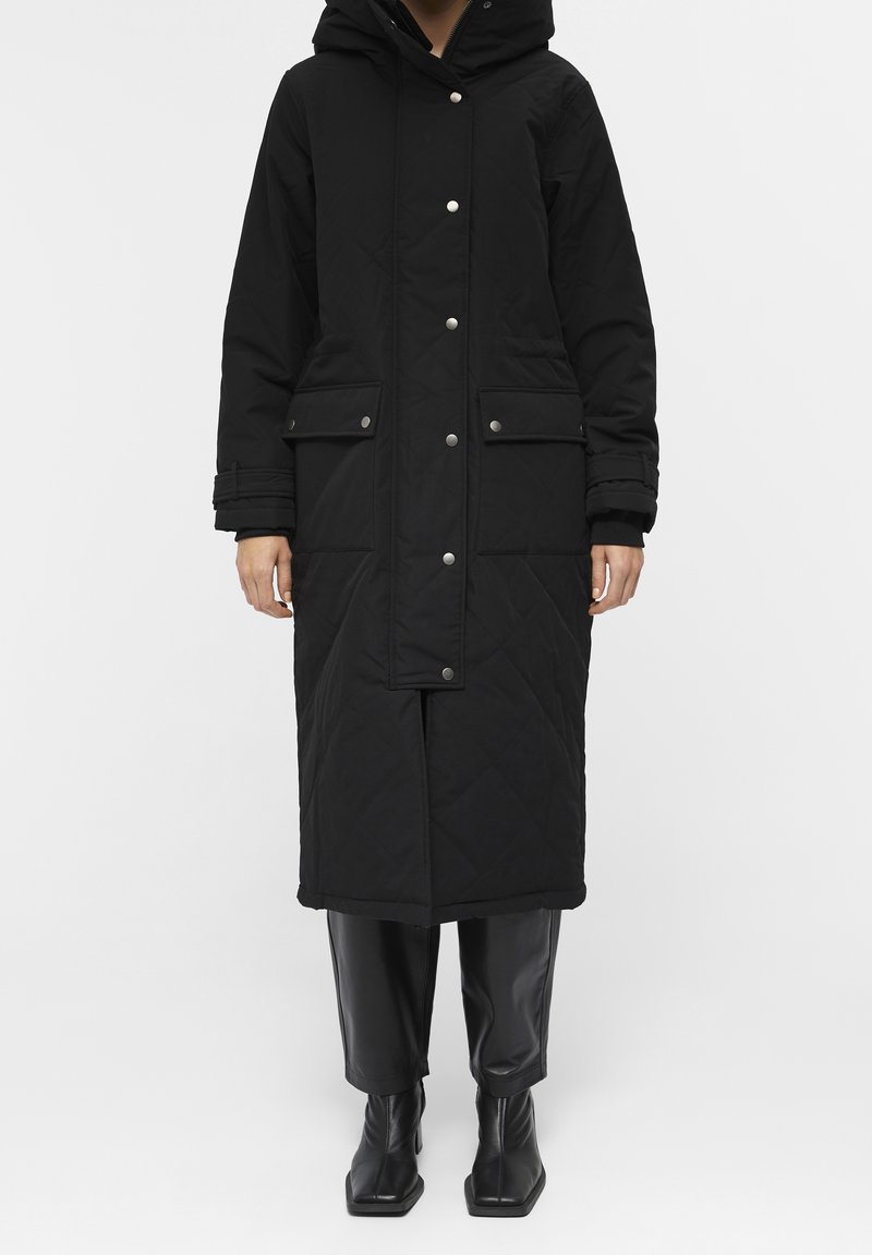 Personne portant un long manteau d'hiver noir à capuche avec des boutons-pression, des poches à rabat, ainsi qu'un pantalon en cuir noir et des bottes visibles sous le manteau.