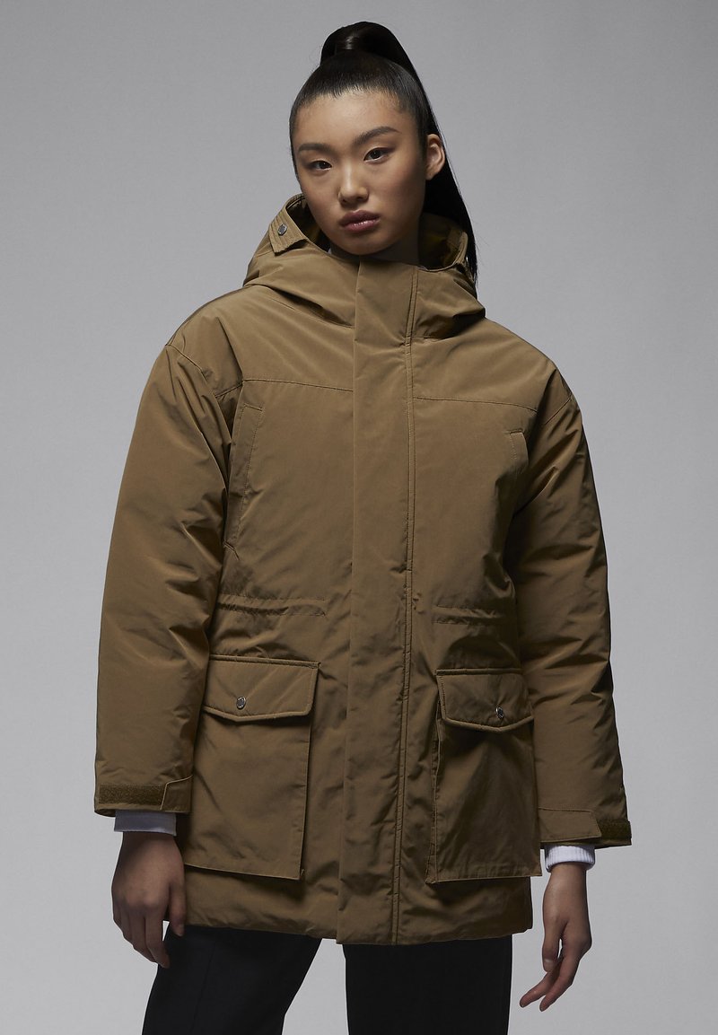 Joven con pelo largo en una coleta alta, vistiendo una chaqueta de invierno marrón de gran tamaño con capucha y grandes bolsillos frontales.