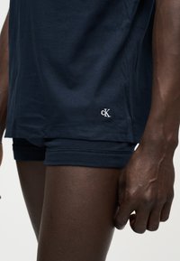 Κοντινό πλάνο σκοτεινόχρωμου ατόμου που φοράει σκούρα μπλε σορτς και αντίστοιχο πάνω μέρος με μικρό λευκό λογότυπο «cK» στη ραφή του πουκάμισου.