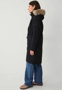 Zwarte parka-jack met een met bont gevoerde capuchon, voorzien van een tailleband met trekkoord, zijzakken en een knoopdetail op de mouw. Draag over losse jeans.