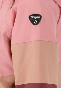 Giacca della marca Zigzag con tessuto rosa e beige, presenta una patch logo nera sulla manica, texture liscia e design a blocchi di colore.