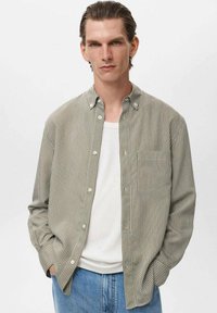 Camisa de manga larga a rayas en verde y blanco, con botones en la parte delantera y un bolsillo en el pecho, llevada sobre una camiseta sin mangas blanca, combinada con jeans azules.