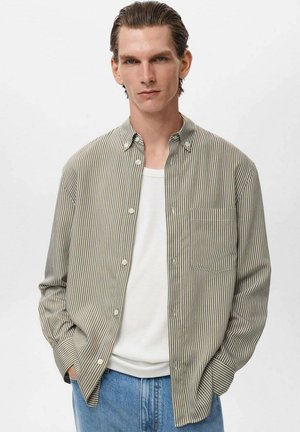 Camicia a righe a maniche lunghe verde e bianca, con bottoni frontali e taschino sul petto, indossata sopra una canotta bianca, abbinata a jeans blu.