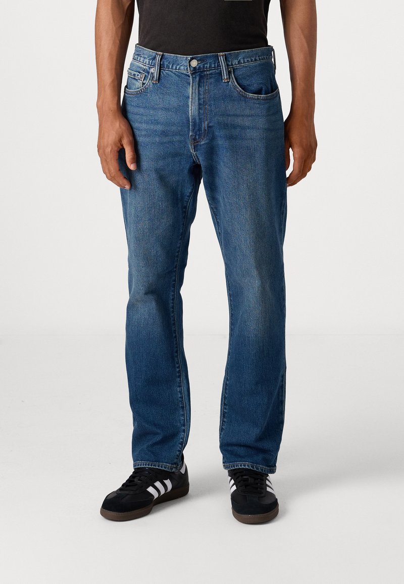 Vaqueros de mezclilla azul con corte recto, cinco bolsillos y cierre de botón. Combinados con zapatillas deportivas negras con franjas blancas.