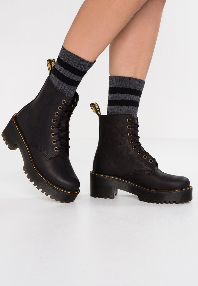 Jambes portant des bottes à plateforme en cuir noir à lacets avec des coutures jaunes et des chaussettes grises à deux rayures noires.