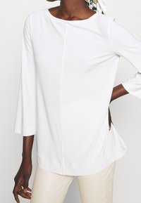 Biała bluzka z okrągłym dekoltem, rękawami 3/4 i subtelnym szwem z przodu. Materiał wydaje się gładki i lekko teksturowany.