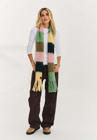 Kobieta stojąca, ubrana w długi, wielokolorowy szalik w paski, białą koszulę z długim rękawem, ciemne szerokie spodnie i beżowe buty turystyczne na białym tle.