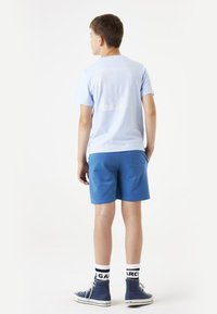T-shirt en coton bleu clair avec un design imprimé dans le dos, associée à un short bleu. L'ensemble est complété par des baskets bleues et des chaussettes à motifs.