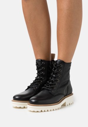 Bottines en cuir noir à bout rond, à lacets sur l'avant, avec une semelle extérieure blanche épaisse. Elles présentent une finition texturée et un accent beige au niveau du talon.