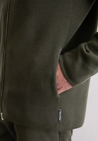 Olive green knit sweater with textured fabric, front zipper, and side pockets. Features a fabric tag at the hem.