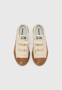 Beige lerretsneakers med brune gummitåhette, borrelåsremmer og teksturerte brune gummisåler. Har "Converse All Star" merke.