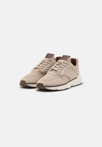 Beige athletic shoes with a textured knit upper, lace-up design, brown accents on the tongue, and a contrasting white midsole.