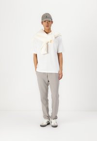 Weißes T-Shirt mit kurzen Ärmeln, hellgraue Jogginghose, cremefarbener Pullover um die Schultern gebunden, graue Mütze und schwarz-weiße Sneakers. Ein einfacher, lässiger Look.